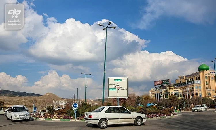 میدانی در محله حکیمیه با مسجد گنبدسبز، تابلوی مسیر و تپه‌های شرق تهران زیر آسمان ابری