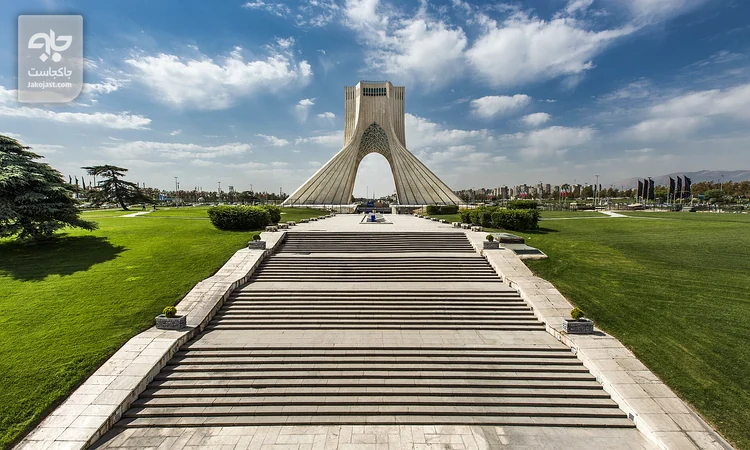 برج آزادی تهران با پلههای سنگی، چمن سبز و آسمان ابری بهاری در میدان آزادی