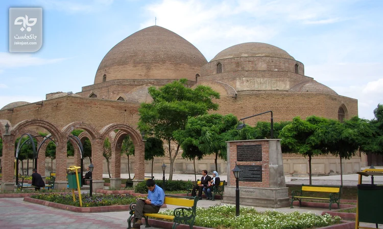 نمای بیرونی مسجد کبود تبریز با دو گنبد آجری، رواق‌های قوسی و فضای سبز جلوی بنا